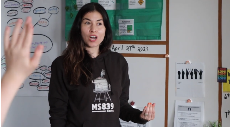 The image shows a woman in a classroom setting. She is wearing a black hoodie and appears to be interacting with someone off-camera, possibly a student, as she raises her hand. The background includes a whiteboard with writing and drawings, suggesting an educational environment. The date "April 27th 2023" is visible on the wall.